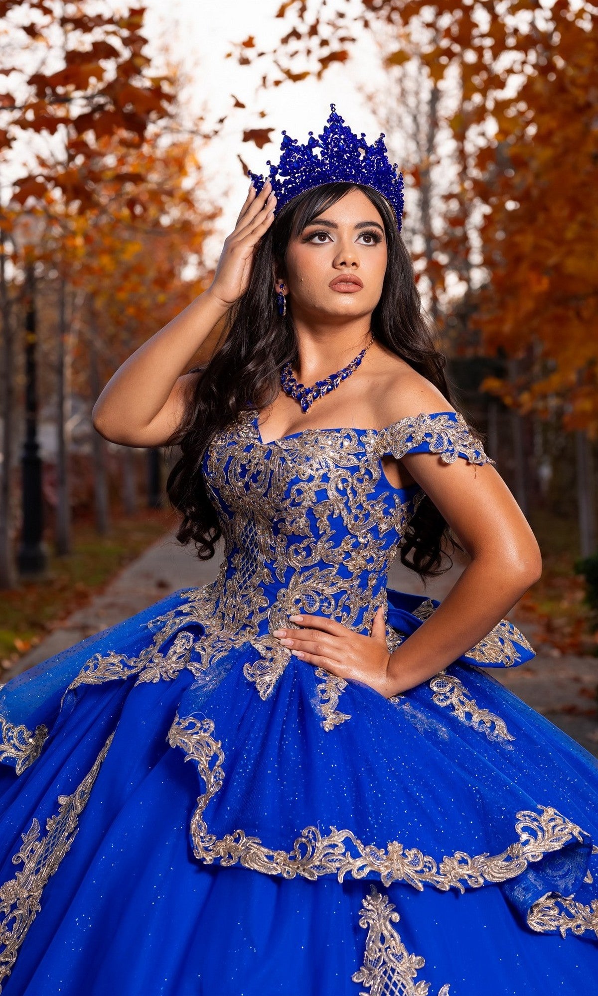 Wearing the Quinceanera Dress CHF3120 By Chicas, a woman crowned and confident poses on a leaf-covered path, surrounded by autumn trees.